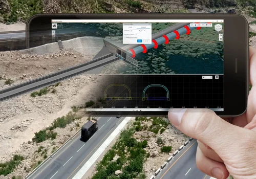 A person’s hand holding a smartphone with the SYNCHRO Digital Construction Software, displaying a 3D modeling app that overlays a digital barrier on a real roadway beside a river. A person's hand holding a smartphone with the SYNCHRO Digital Construction Software, displaying a 3D modeling app that overlays a digital barrier on a real roadway beside a river.
