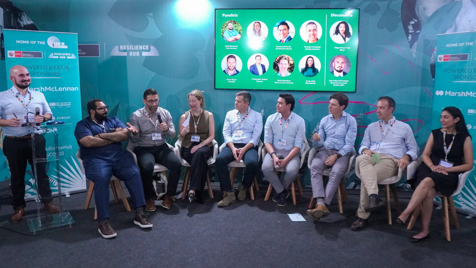 Nine people sit on chairs in a panel discussion, with microphones and badges, facing an audience. A screen behind them displays panelist photos and discussion topics. | Bentley Systems | Infrastructure Engineering Software Company Nine people sit on chairs in a panel discussion, with microphones and badges, facing an audience. A screen behind them displays panelist photos and discussion topics.
