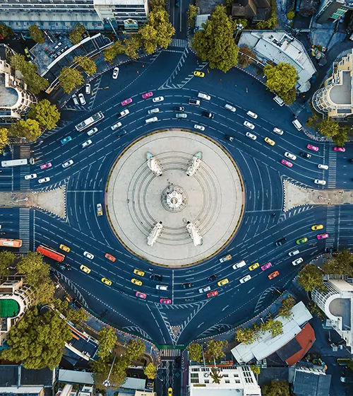 Aerial view of a large roundabout with multiple lanes of cars surrounding a central monument, bordered by trees and buildings.