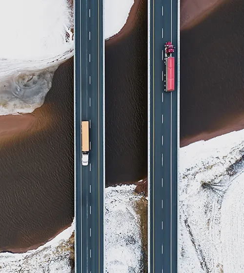 Aerial view of two parallel highways with large trucks traveling in opposite directions, surrounded by snowy and brown terrain.