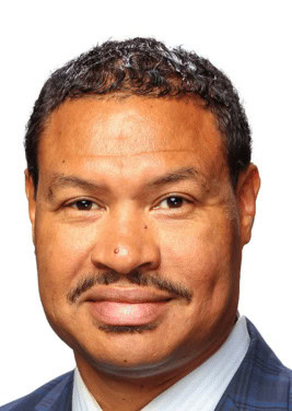 A person with short curly hair, a mustache, and a goatee, wearing a pinstripe suit and light blue shirt, stands against a white background. A natural blend of design elegance and structural precision is evident in their poised appearance.