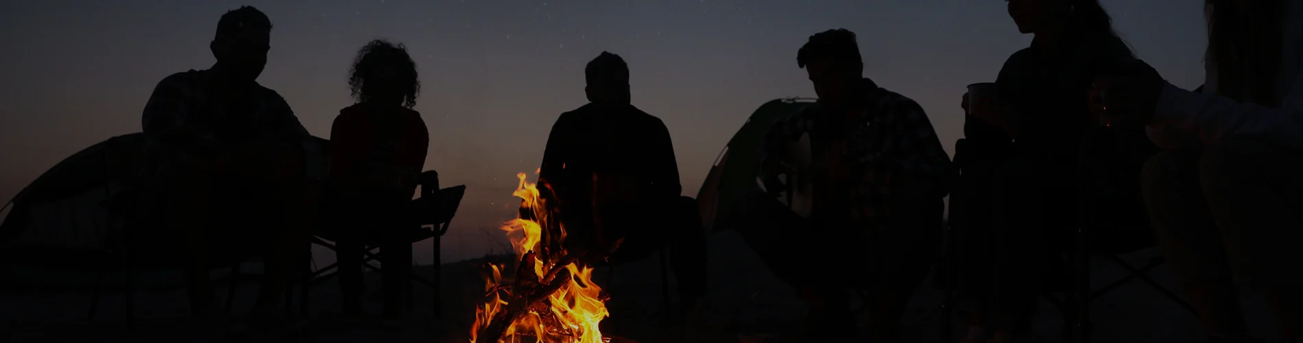 A group of people sit in a circle around a campfire at night, engaged in Fireside Chats, silhouetted against the dark sky.