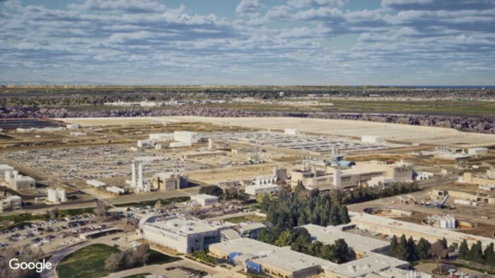 An aerial view of an expansive industrial complex with various buildings and infrastructure. The background features a wide landscape under a partly cloudy sky. The image showcases the integration of photorealistic 3D tiles enabled by iTwin, using Google 3D tiles and Cesium technology.