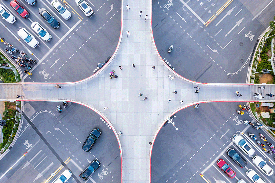 Aerial view of a cross-shaped pedestrian overpass above a busy intersection with cars and people visible on the roads and sidewalks below. | Bentley Systems | Infrastructure Engineering Software Company Aerial view of a cross-shaped pedestrian overpass above a busy intersection with cars and people visible on the roads and sidewalks below.