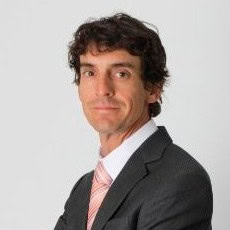 A man with curly hair, leveraging the power of design, poses confidently in a suit and pink tie against a plain white background.