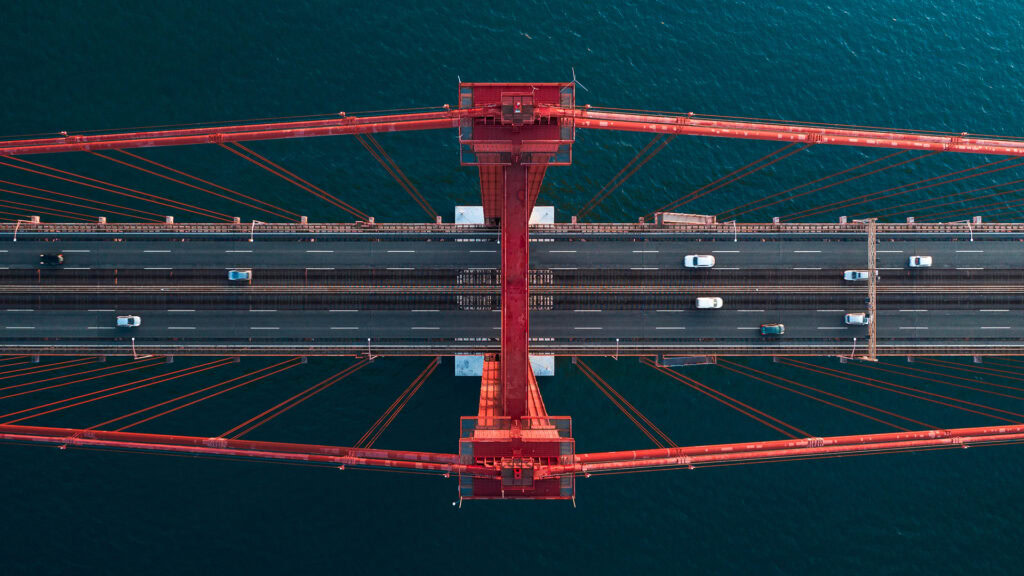 Aerial view of a red suspension bridge in 2024, showcasing the vibrant scene of transportation with multiple vehicles driving on it, set against a backdrop of blue water. | Bentley Systems | Infrastructure Engineering Software Company Aerial view of a red suspension bridge in 2024, showcasing the vibrant scene of transportation with multiple vehicles driving on it, set against a backdrop of blue water.