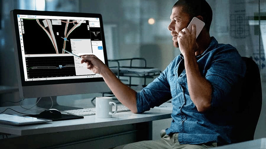 A man sits at a desk talking on a phone while pointing at a computer screen displaying engineering design software, reviewing a COPEL Distribuição case study. | Bentley Systems | Infrastructure Engineering Software Company A man sits at a desk talking on a phone while pointing at a computer screen displaying engineering design software, reviewing a COPEL Distribuição case study.